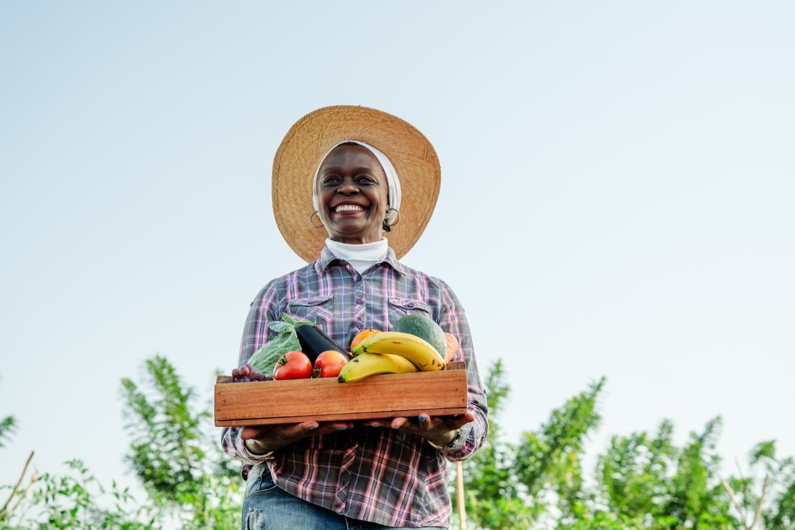 A smiling farmer in a straw hat holds a wooden crate of fresh produce under an open sky.